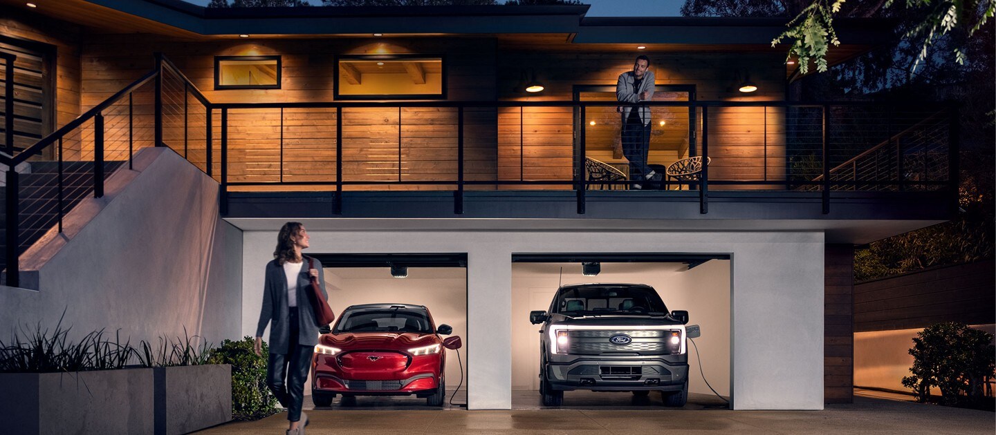 Una Ford Mustang Mach-E® y una Ford F-150® LightningTM 2022 se cargan en un garaje por la noche cerca de dos personas