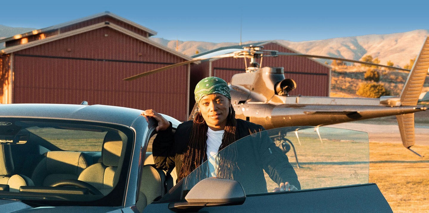 A person standing near the driver-side door of a 2023 Ford Mustang® coupe with a helicopter in the background