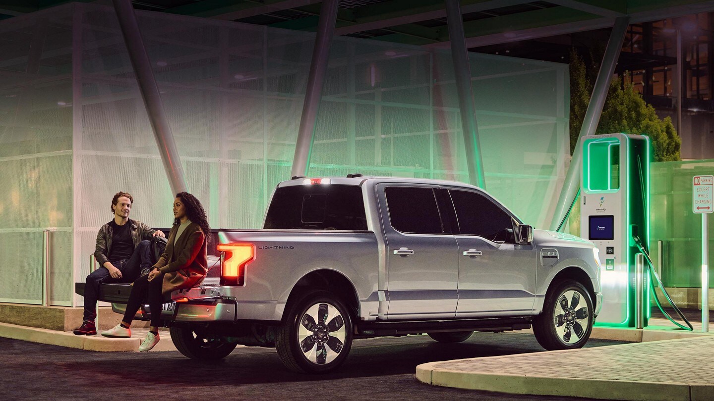 A couple sits on the open liftgate of a silver Ford F-150® Lightning® as it charges