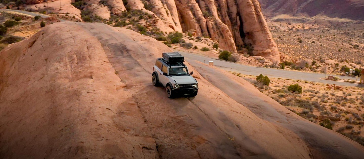 A gray two-door 2023 Ford Bronco® drives down a rock formation with a cargo carrier attached to its roof