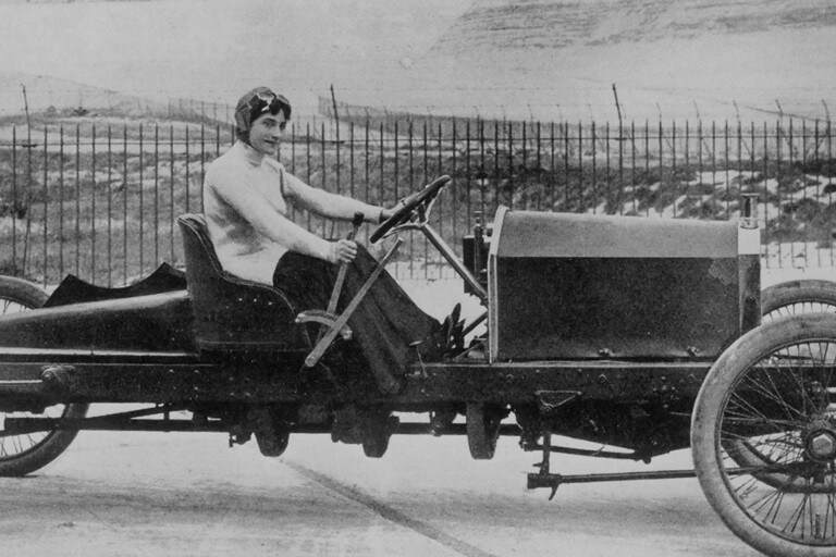 We see a black and white portrait of auto innovator Dorothy Levitt driving a 1920s speedster wearing a helmet with goggles tucked on top of it.