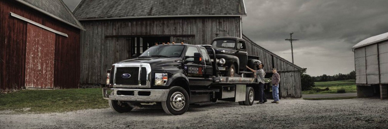 Two men loading a classic [insert model if desired] car onto the bed of a Ford tractor trailer in a farmyard