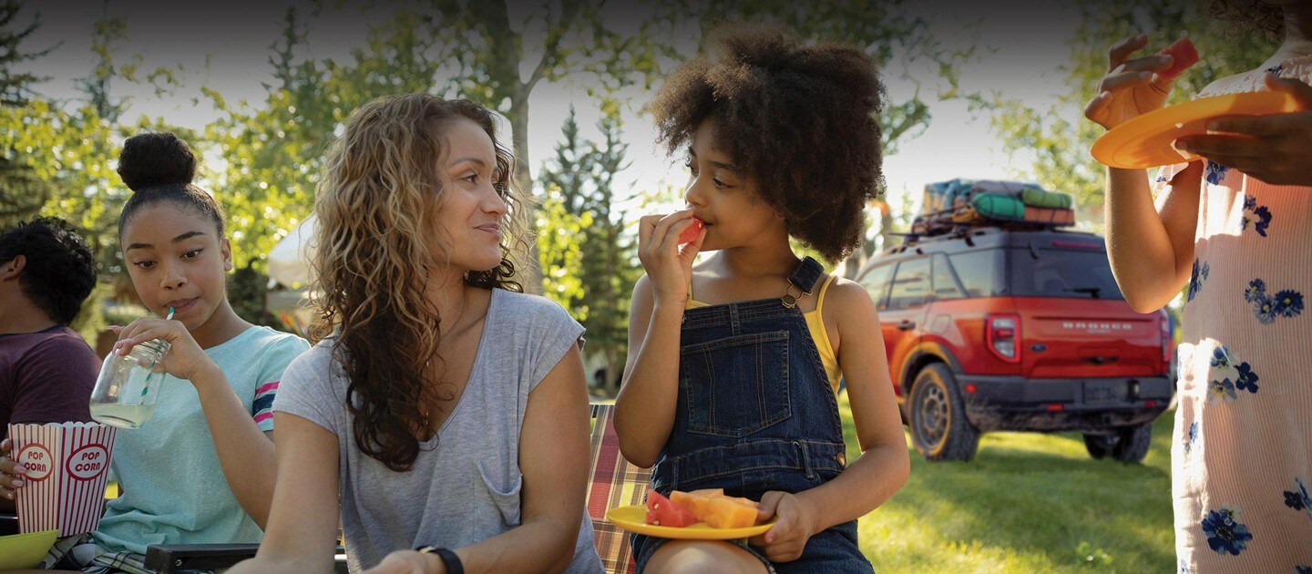 A woman and two young girls have a picnic with a 2023 Ford Bronco in the background