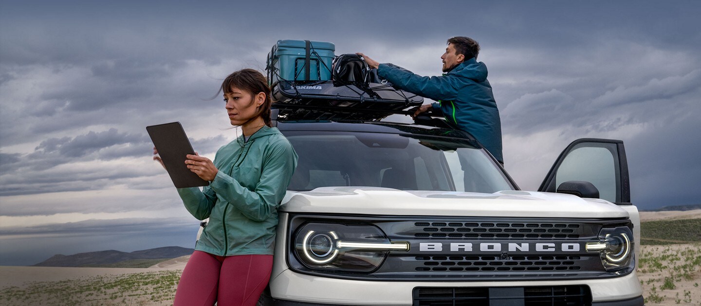 A woman leans back on a 2022 Ford Bronco® Sport SUV, while a man secures cargo to the roof of the vehicle.