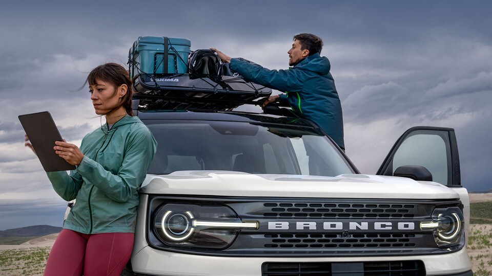 A woman leans back on a 2022 Ford Bronco® Sport SUV, while a man secures cargo to the roof of the vehicle.