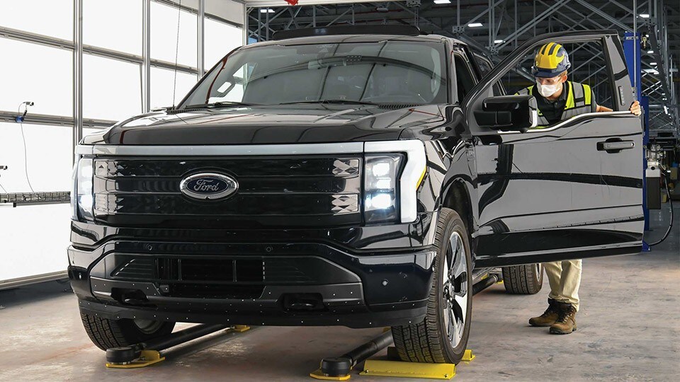 Ford factory worker in yellow hard hat and mask inspecting black Ford F-150® Lightning™ as it comes off the line