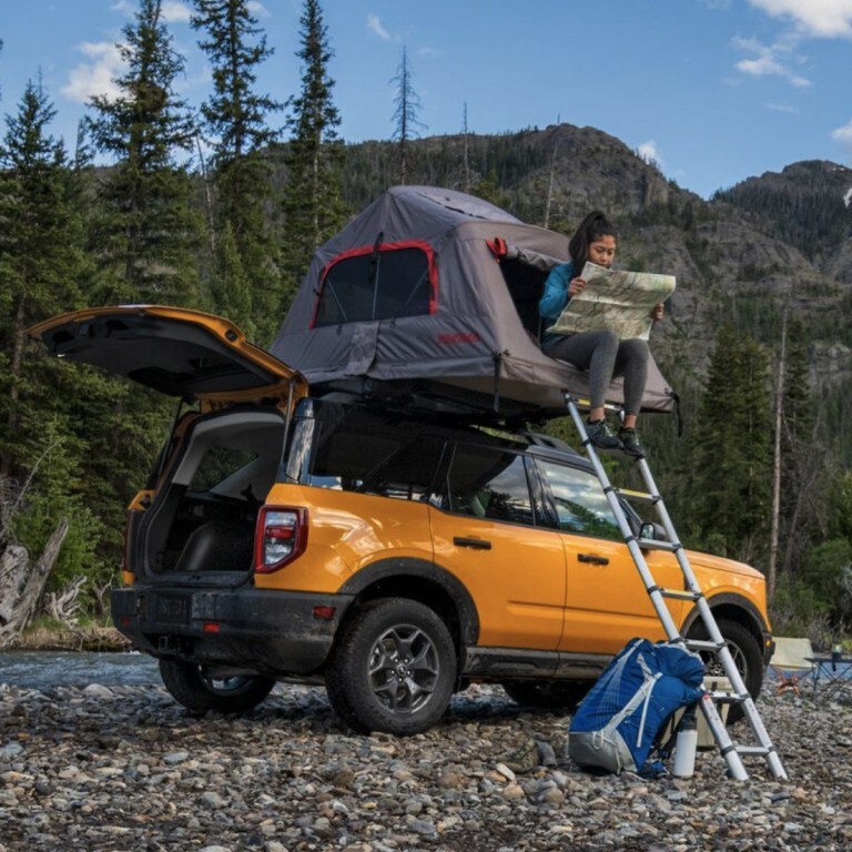 A person is reading a map while sitting in a tent attached to the roof of a Ford Bronco.