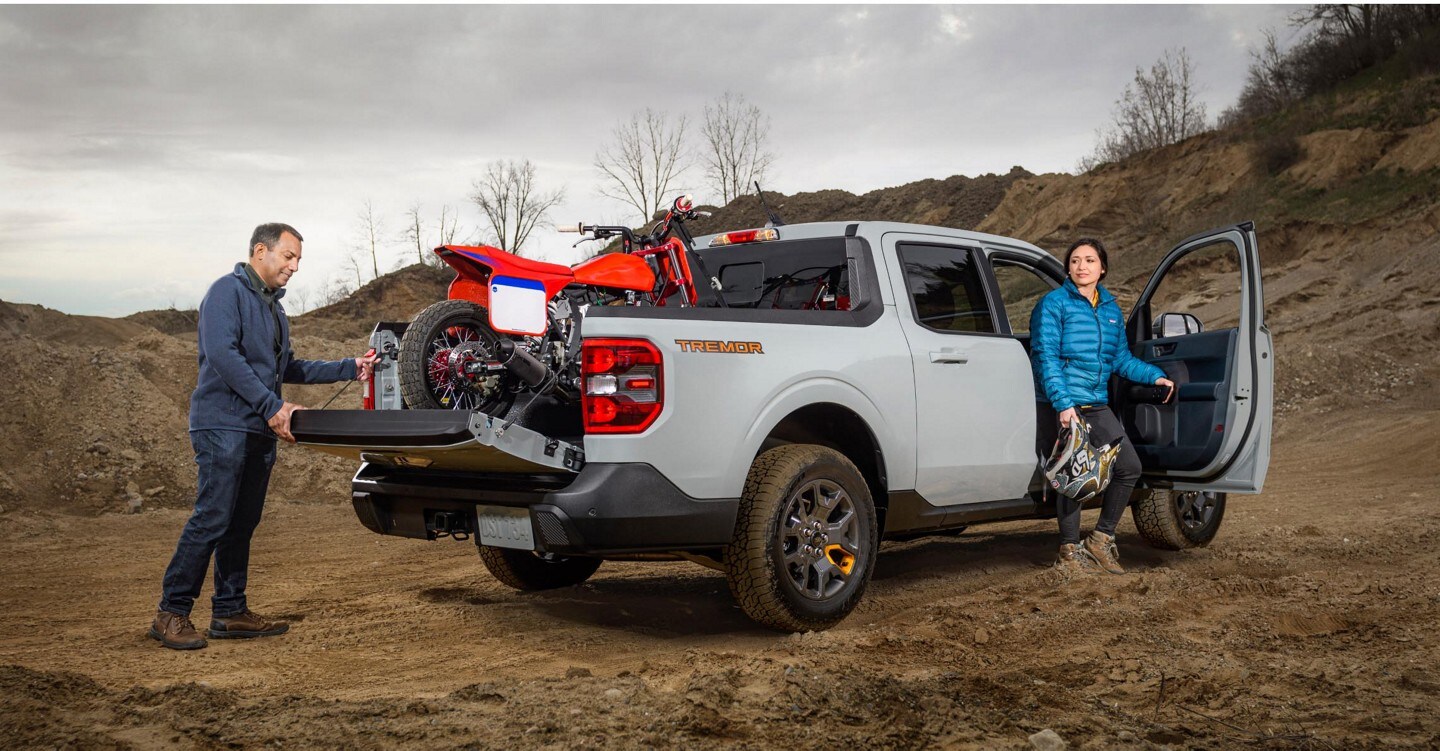 Image of a man lowering the tailgate of a 2023 Ford Maverick™ Tremor® with a motorcycle in the bed of the truck and a woman exiting the passenger side