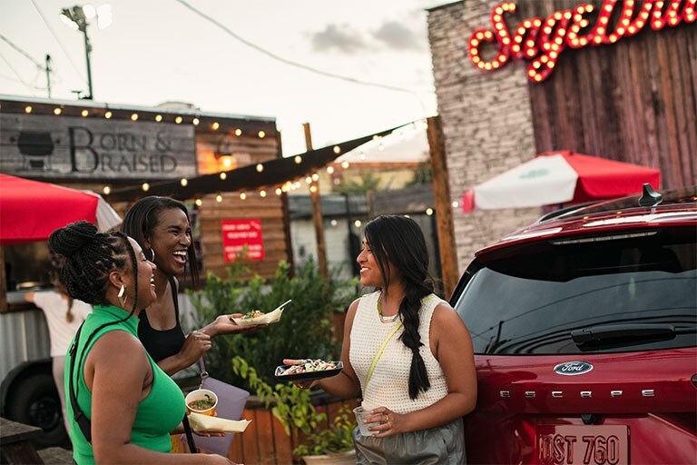 Three people are seen laughing next to a red Ford Escape.