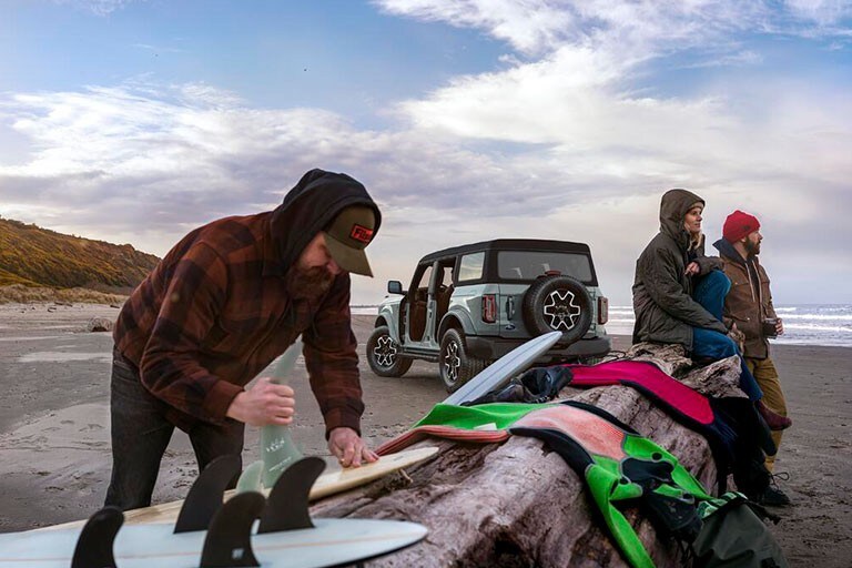 Photo of a group of friends preparing surf boards by a Ford Bronco on the beach.