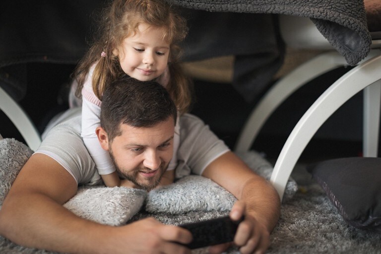 Foto de un padre recostado, mirando el teléfono con un niño sobre los hombros