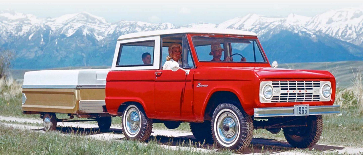 Una familia en un Ford Bronco® Sport Wagon 1966 en Royal Maroon con techo en Wimbledon White remolcando una caravana