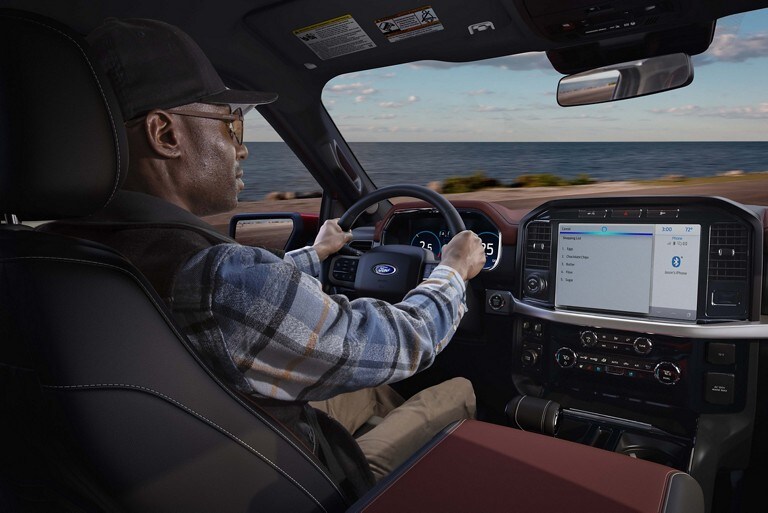 A person behind a wheel driving a 2022 Ford F-150 with the ocean in the background
