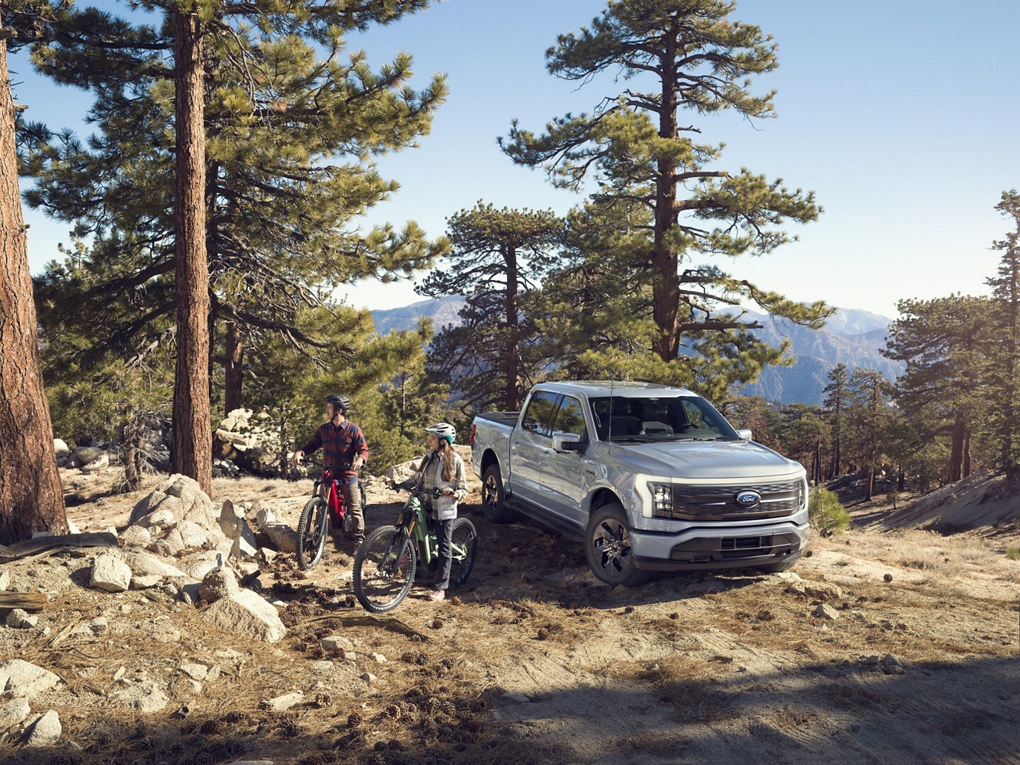 Two cyclists beside an Iced Blue Silver 2022 F-150 Lightning in a mountain forest clearing.