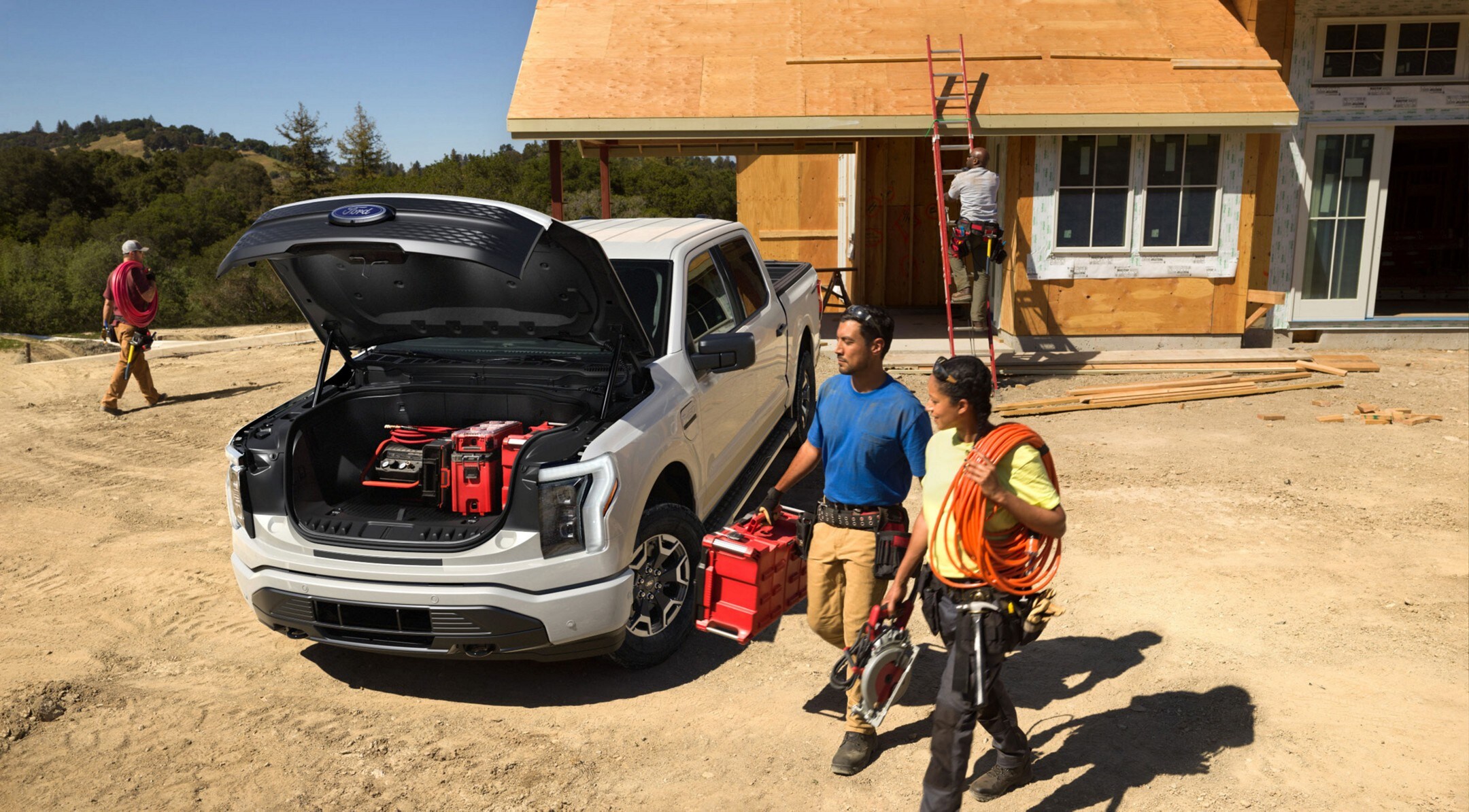Two people at a construction site carrying equipment to the open Mega Power Frunk of an Oxford White 2022 F-150 Lightning