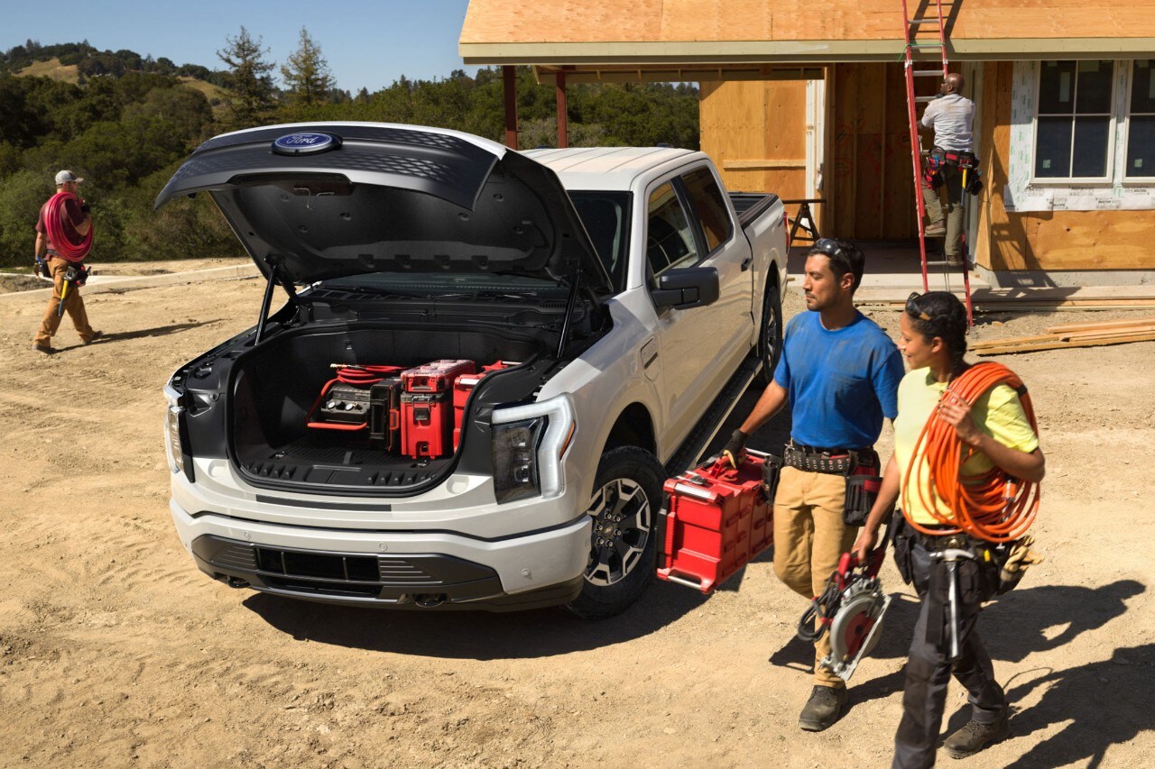 Two people at a construction site carrying equipment to the open Mega Power Frunk of an Oxford White 2022 F-150 Lightning