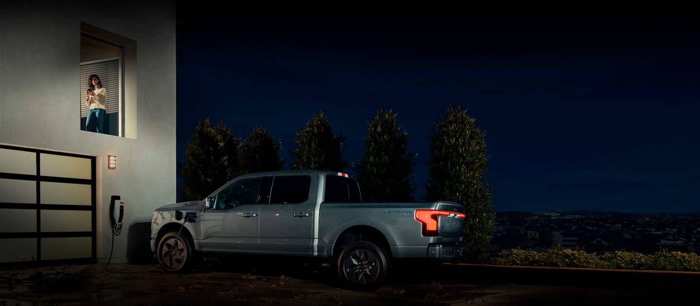 Una mujer consulta la aplicación para smartphone Ford Intelligent Backup Power, mientras una Ford F-150® Lightning™ 2023 está enchufada en su casa
