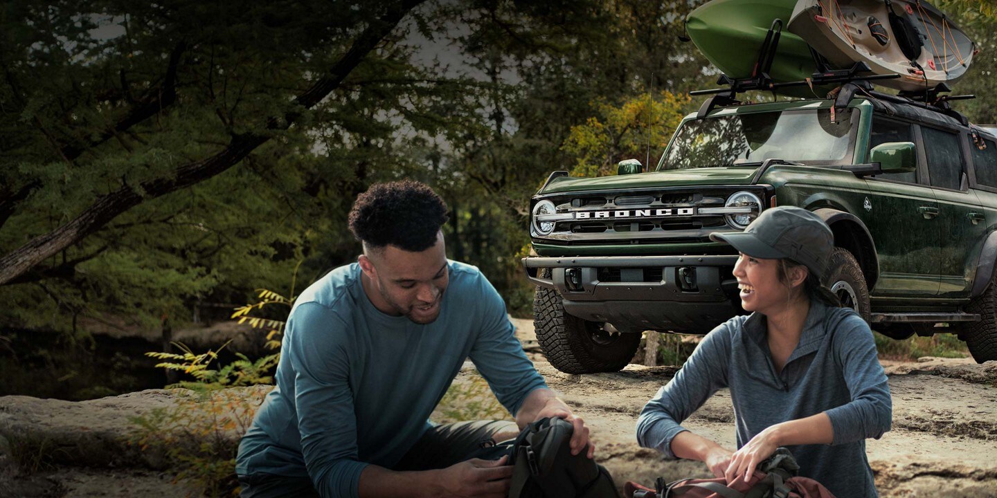 A couple setting up camp with a 2023 Ford Bronco® SUV in background with kayaks on the roof