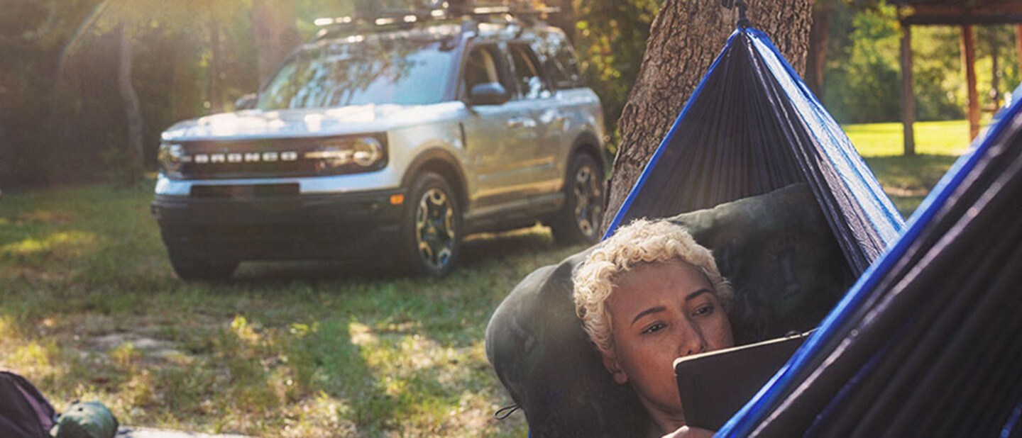 A lady laying in a hammock with gear on a blanket behind her with the vehicle in the background