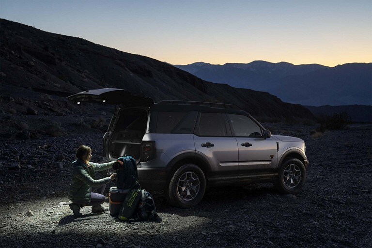 Una persona con equipo de campamento cerca de la puerta trasera levadiza abierta de un modelo Ford Bronco® Sport 2023 con reflectores traseros iluminando