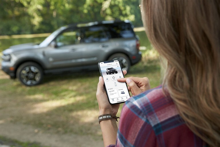 Una mujer mirando la Aplicación FordPass® en su teléfono móvil con la SUV Ford Bronco® Sport 2023 en segundo plano