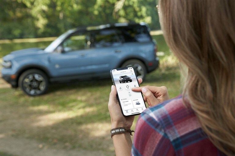 Woman looking at FordPass® App on mobile phone with 2024 Ford Bronco® Sport SUV in the background