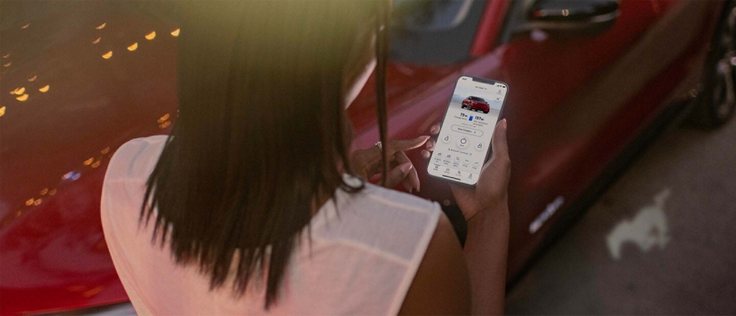 Woman viewing FordPass home screen in front of a Red 2023 Ford Mustang Mach-E
