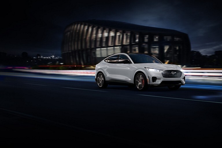 Space White Metallic 2023 Ford Mustang Mach-E® SUV being driven on a highway past a stadium at night