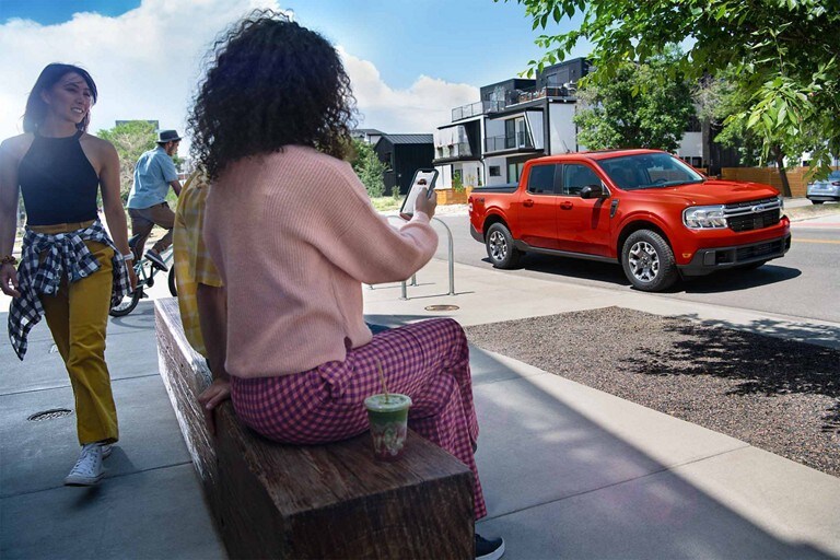 A person is sitting on a bench looking at their phone with a 2024 Ford Maverick® truck parked on a street