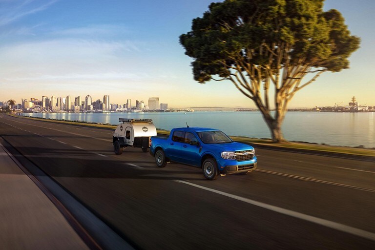 Una camioneta Ford Maverick® LARIAT® 2024 en Atlas Blue Metallic andando con un remolque en una carretera metropolitana cerca del agua