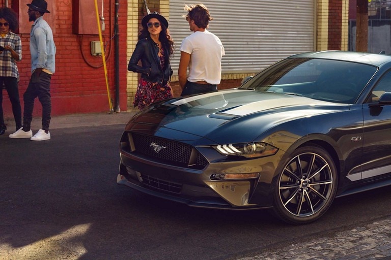 Un Ford Mustang® Coupe 2023 en Carbonized Gray Metallic estacionada en una calle con cuatro personas de pie cerca