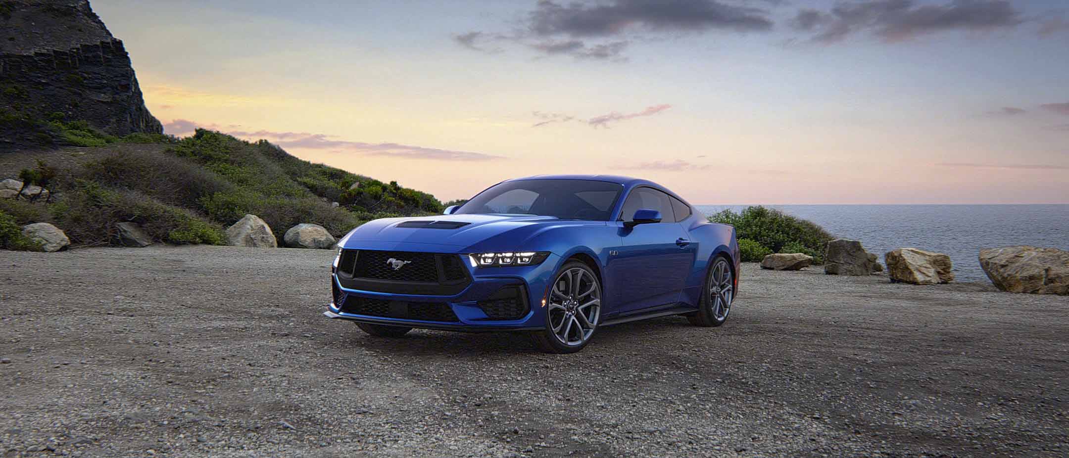 Blue 2024 Ford Mustang parked near ocean with sunset and mountain in background