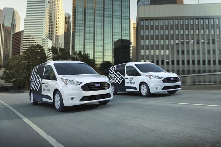 Dos Ford Transit Connect Cargo Van 2023 en Frozen White con gráficos comerciales no originales andando por una calle de ciudad