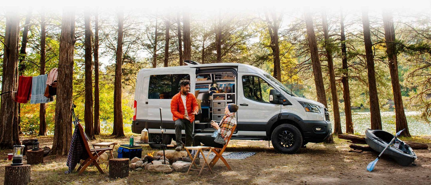 A 2023 Ford Transit Trail™ van parked in the woods next to people having a campfire
