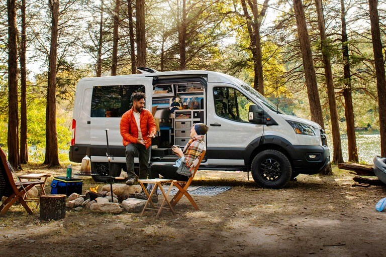 Una Ford Transit Trail™ Van 2023 estacionada en el bosque al lado de personas compartiendo una fogata