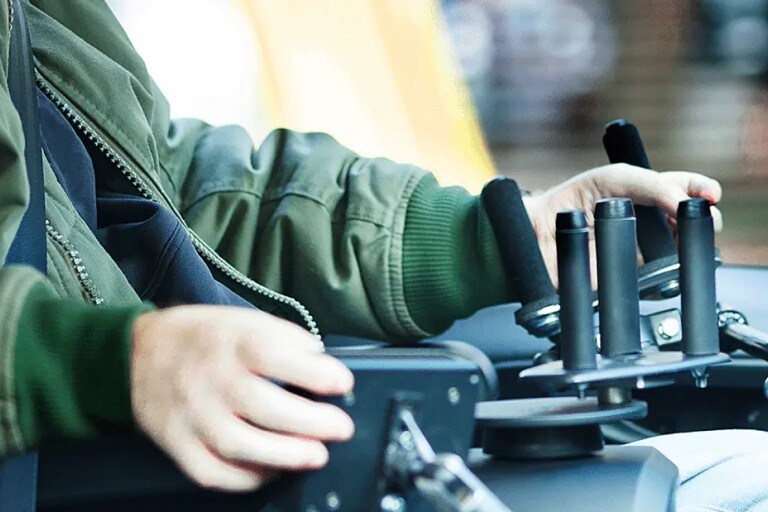 A man is seated in the driver's seat of a vehicle with hand controls installed. He is interacting with one of the controls to his lower right