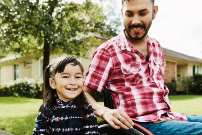 A bearded man in a red and white checked shirt is seated in a wheelchair next to his daughter, who is holding a stack of books