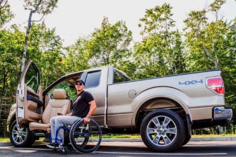 A man wearing a t-shirt, baseball hat and jeans in a wheelchair next to a Ford F-150. The driver's side door is open and a power assist seat is lowered.