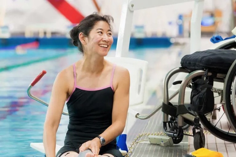 A woman in a bathing suit sitting on the pool deck next to her wheelchair.