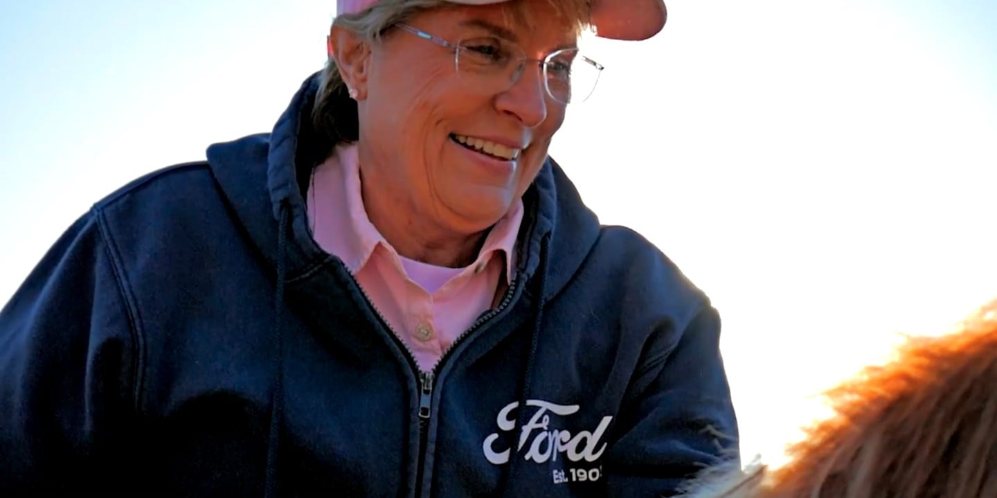 A woman is smiling. She is wearing a cap, glasses and a blue jacket with a Ford name on it.