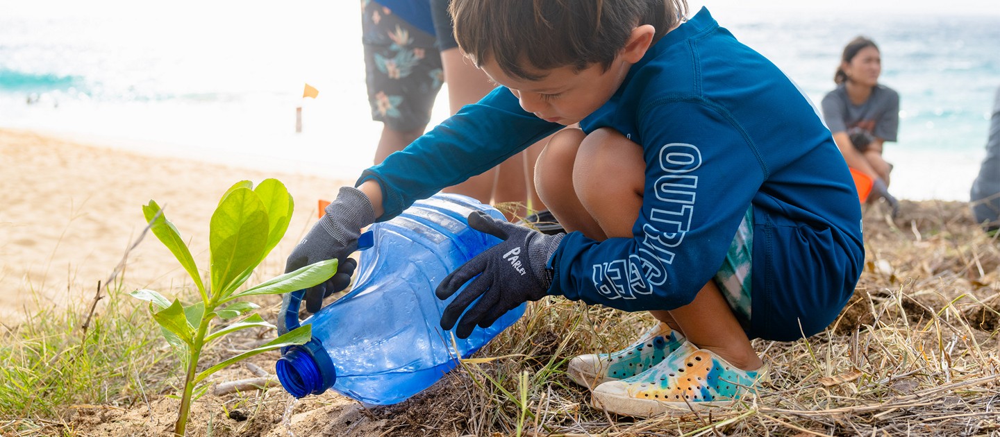 A child watering a plant near the ocean.