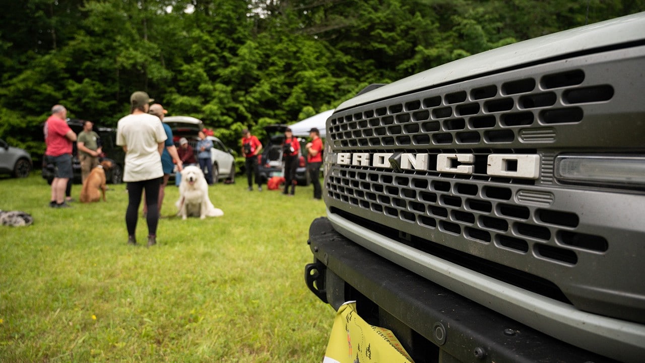 Front bumper of a Bronco® SUV with grant volunteers meeting in a field in the background
