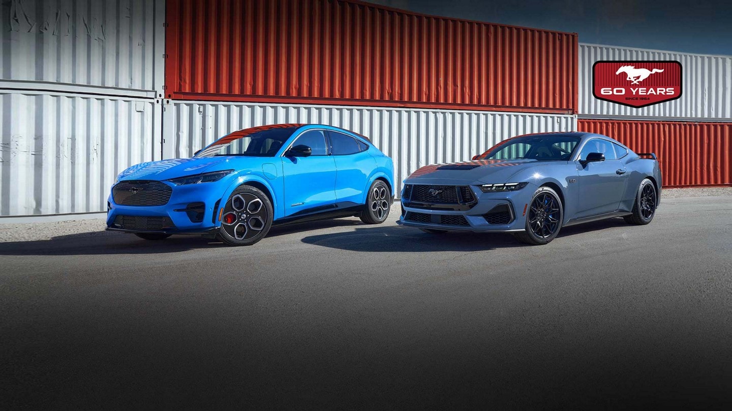 A blue Ford Mustang Mach-E SUV and a gray Ford Mustang Dark Horse coupe parked next to each other in front of a wall of shipping containers