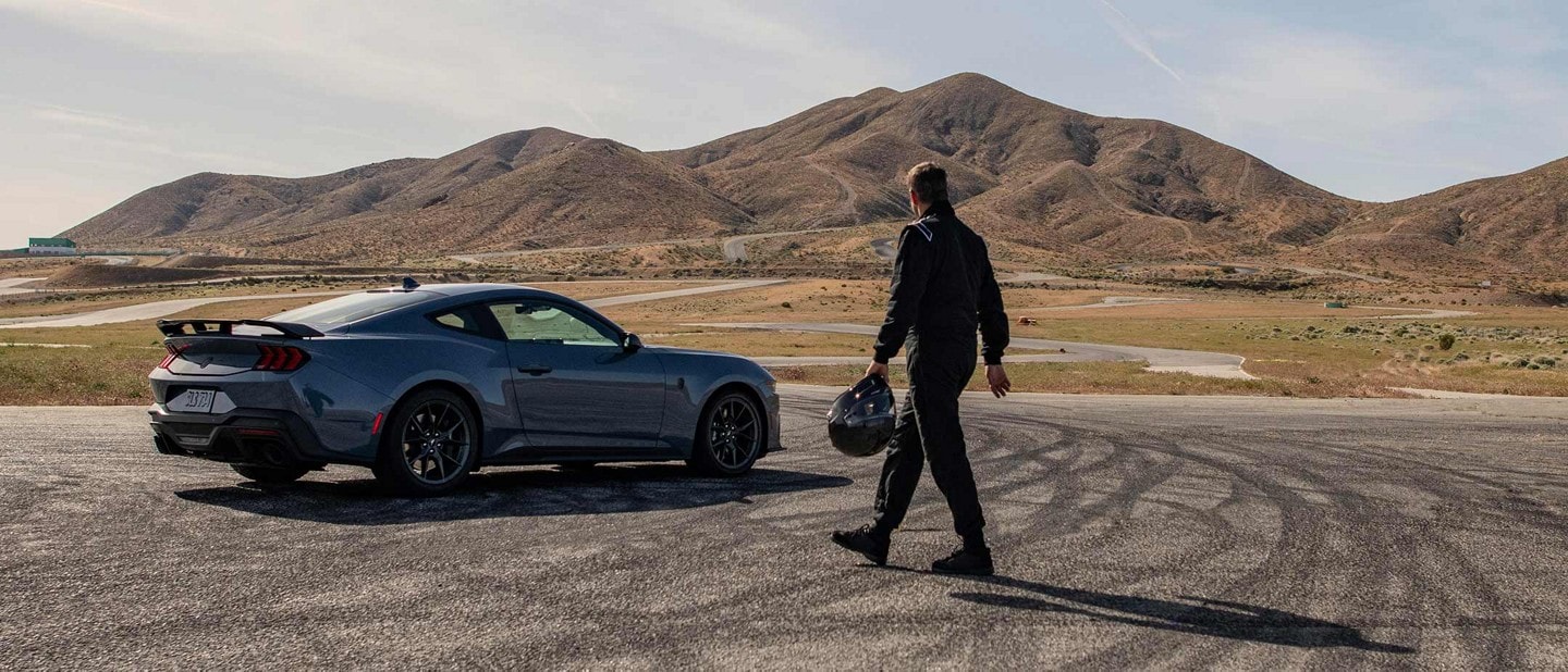 A man wearing racing gear and carrying a helmet walking across an expanse of skidmark-covered pavement toward a parked Mustang Dark Horse coupe