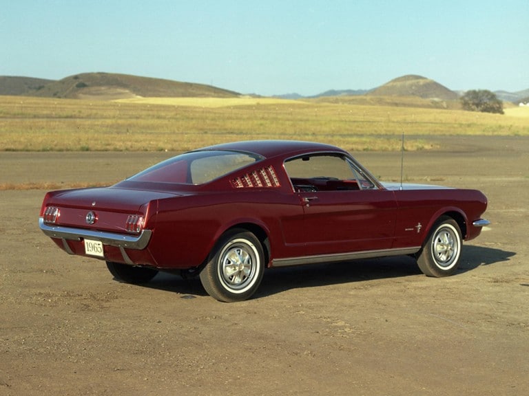 A red 1965 Mustang Fastback parked on a road near a rural landscape