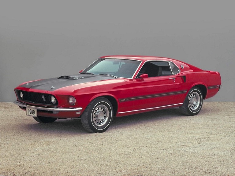 A 1969 Mustang Mach 1, red with a black hood and racing stripes, parked on an expanse of gravel in front of a blank gray wall