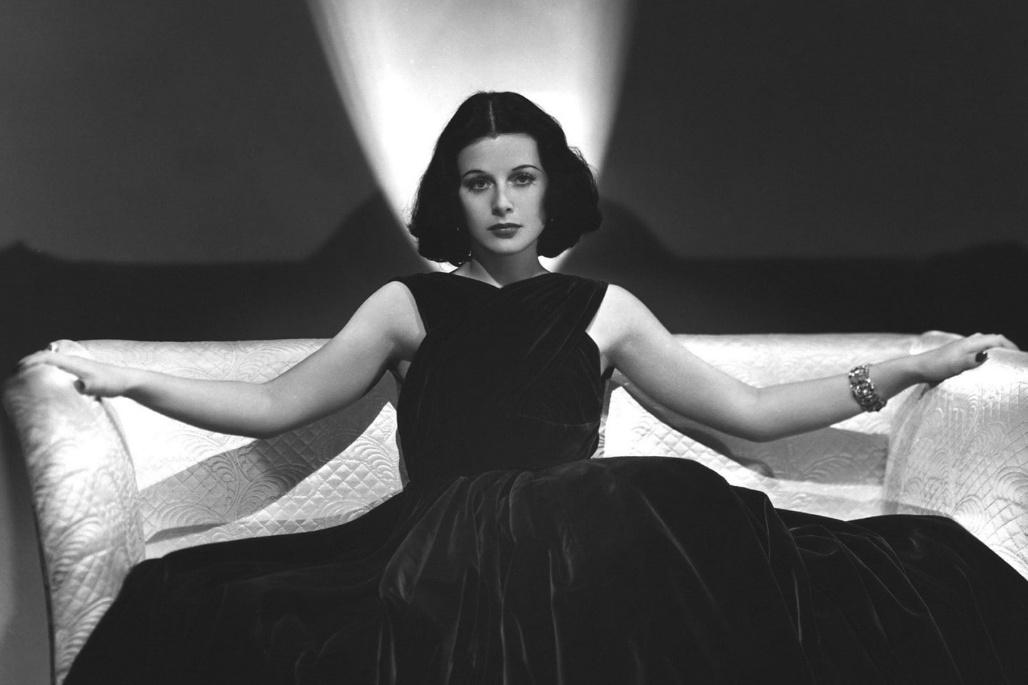 We see a black and white photo of actress and inventor Hedy Lamarr in a black dress sitting on a sofa with a spotlight behind her. At bottom of her photo, we see the words “Hedy Lamarr. Glamorous actress. Brilliant inventor.”