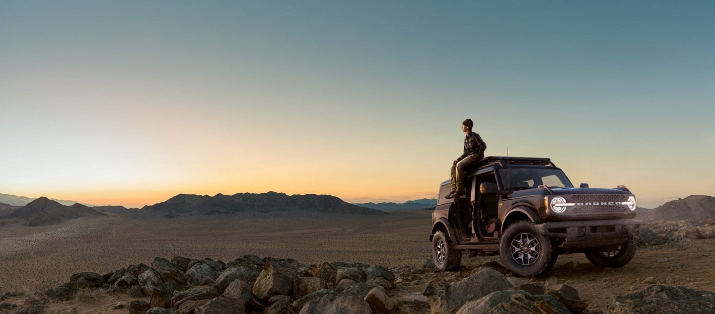 A person sitting on top of a 2021 Ford Bronco parked in the desert at sunrise