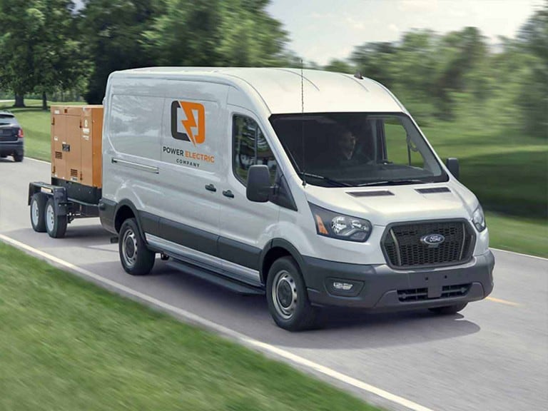 A white 2025 Ford Transit® van pulling a trailer down a road through a grassy park
