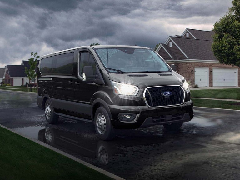 A black 2025 Ford Transit® van driving down a suburban street during a rainstorm